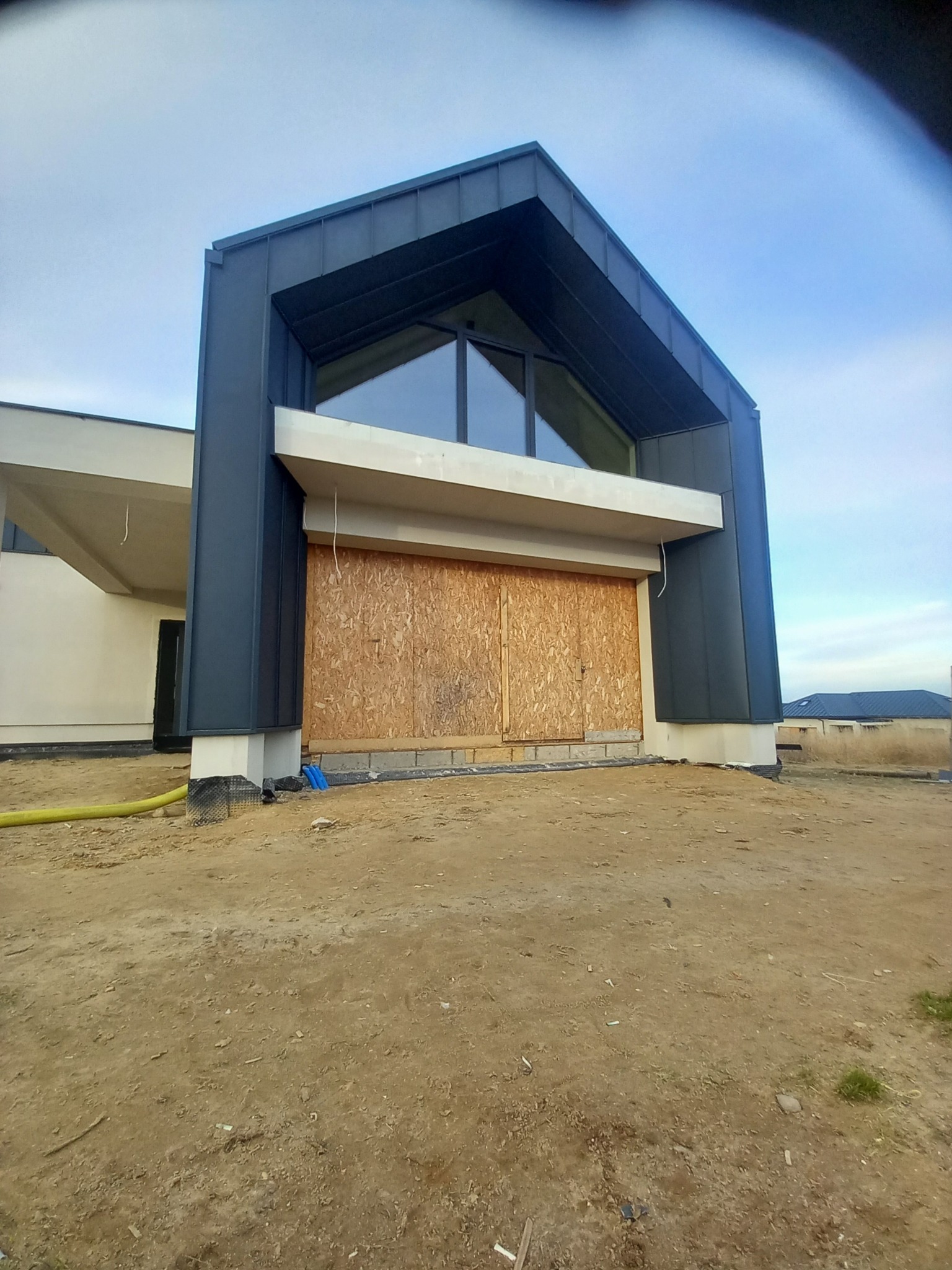 Nowoczesny dom w trakcie budowy z ciemną, metalową obudową dachu i deskami zakrywającymi otwór garażowy.