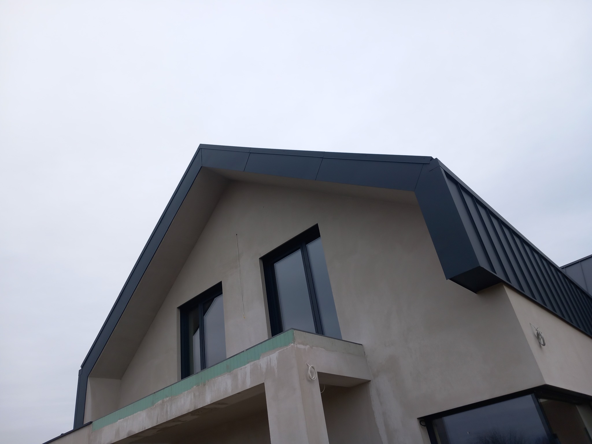 Fragment dachu z ciemnej blachy na nowo budowanym domu z widocznymi oknami i elementami konstrukcyjnymi balkonu, na tle pochmurnego nieba.