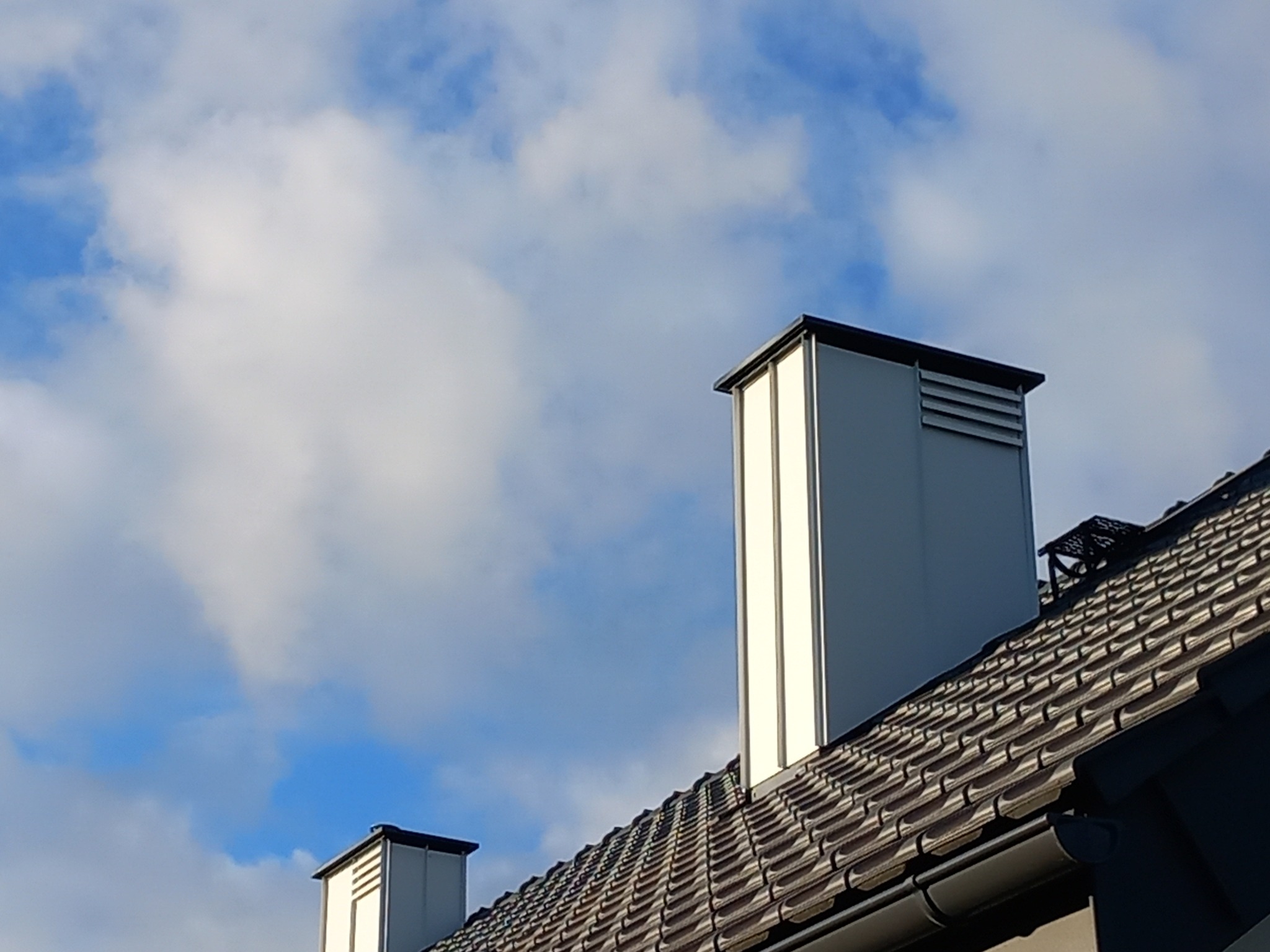 Nowoczesny dom z dachówką ceramiczną i metalowymi kominami wentylacyjnymi na tle błękitnego nieba z białymi chmurami.