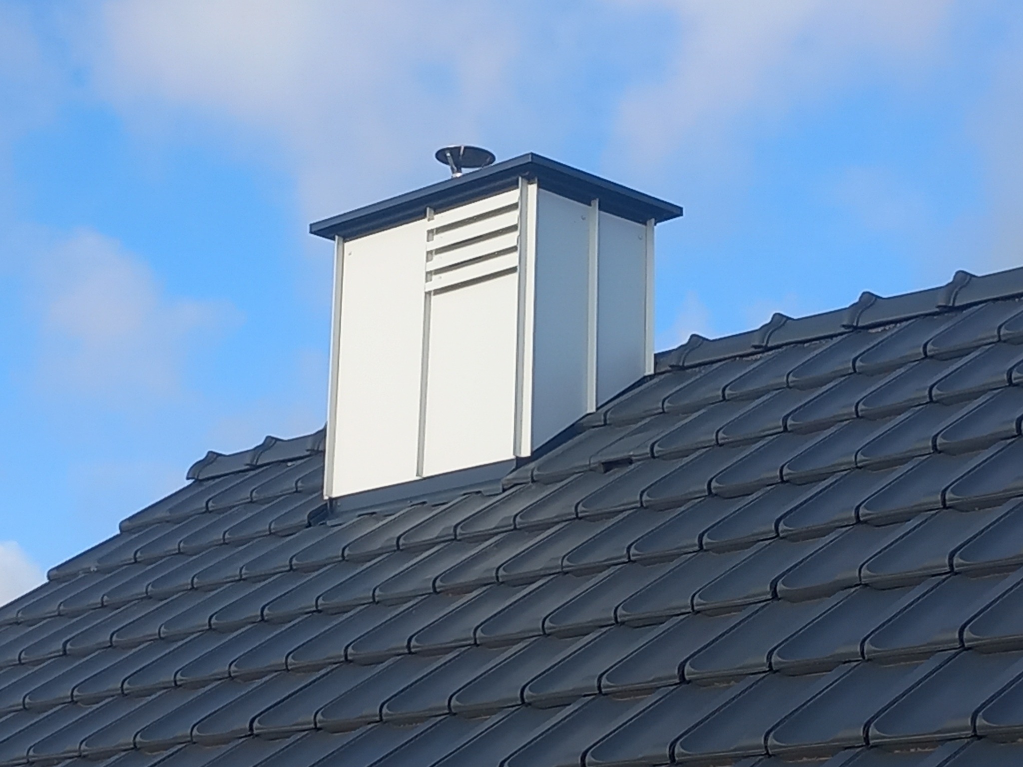 Szczegółowe ujęcie dachu z ciemnej dachówki ceramicznej i białego komina z czarną obwódką na tle błękitnego nieba.