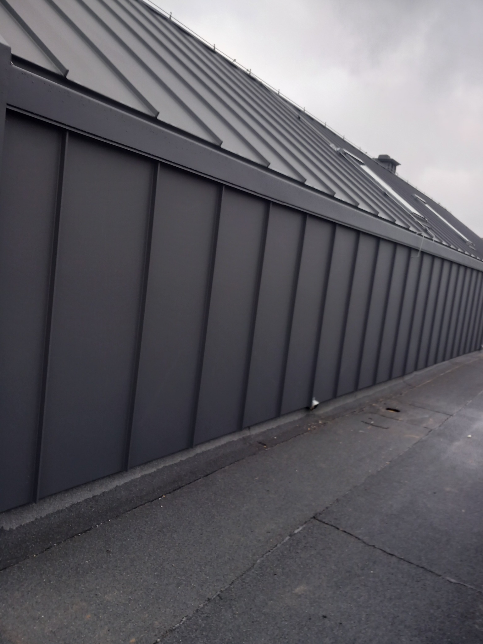 Szary dach z blachy na rąbek stojący widziany z dołu, z widocznym fragmentem dachu płaskiego i pochmurnym niebem w tle.