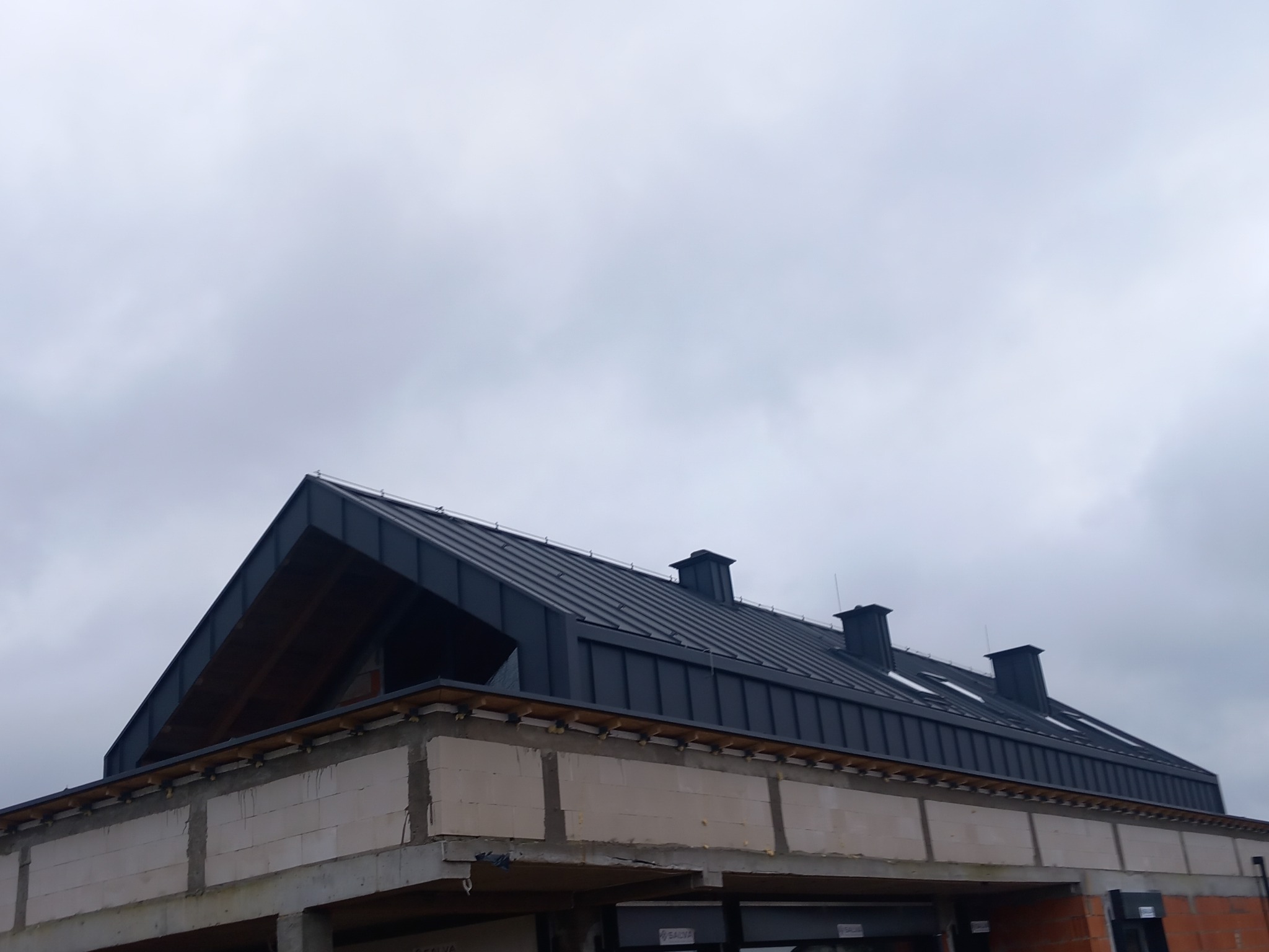 Nowoczesny, ciemny dach z blachy na budynku w trakcie budowy, widoczne kominy i okna dachowe, pochmurne niebo w tle.