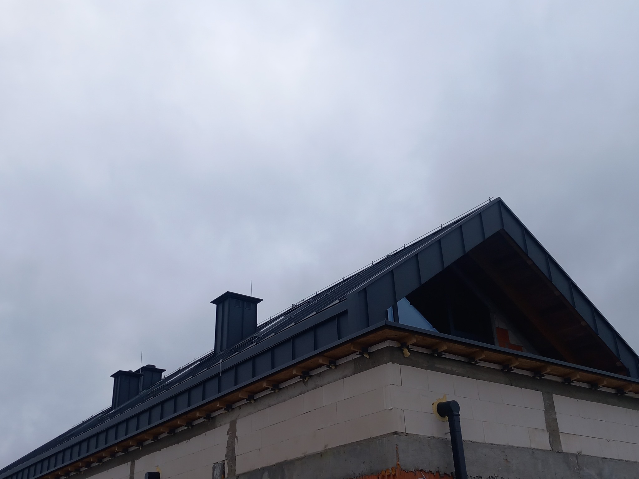 Nowoczesny dom w trakcie budowy, z ciemnoszarą blachą na dachu i widocznymi kominami na tle pochmurnego nieba.