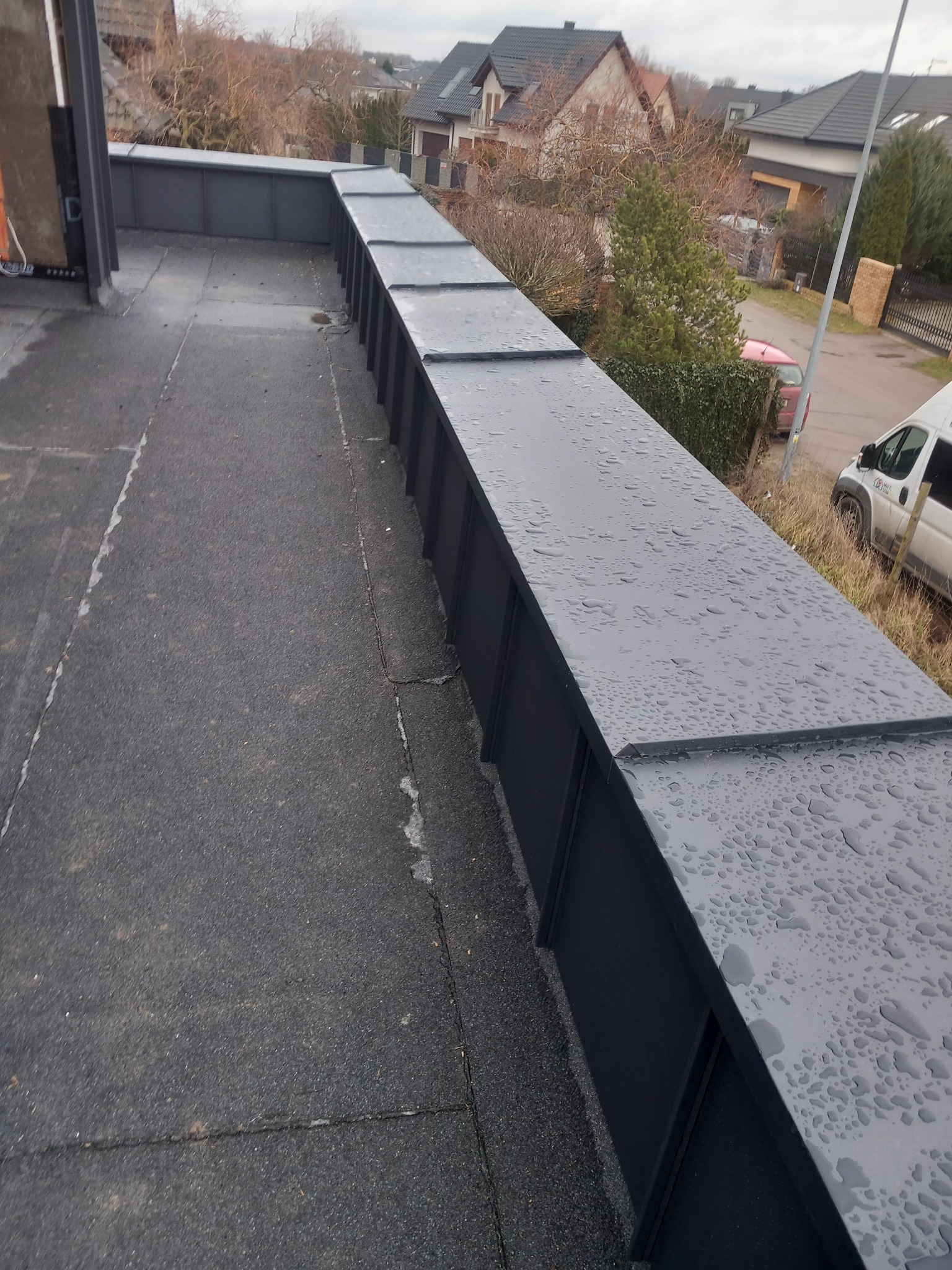 Widok na płaski dach pokryty papą, z zamontowaną obróbką blacharską w kolorze grafitowym, na której widoczne są krople deszczu. W tle zabudowa jednorodzinna i pochmurne niebo.