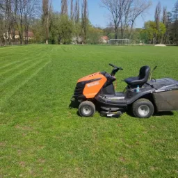 Traktorek do koszenia trawy marki Oleo-Mac na tle boiska z równo przystrzyżoną trawą w pasy.
