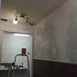 Ściana w trakcie prac przygotowawczych: częściowo zagruntowana, z widocznymi śladami szpachlowania i nierównościami, dolna część wyłożona ciemnymi płytkami. W tle drabina i fragment pomieszczenia...