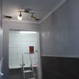 Świeżo pomalowane szare ściany w pomieszczeniu z widocznym fragmentem łazienki z białą glazurą i drabiną, oświetlenie sufitowe z trzema reflektorami.