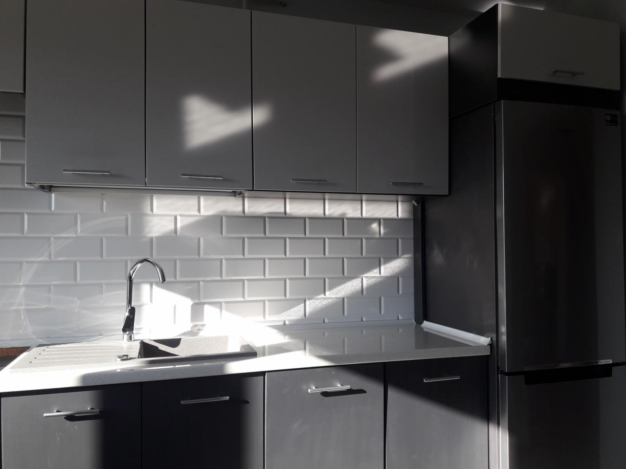 Nowoczesna kuchnia z szarymi szafkami, białymi płytkami na ścianie i promieniem słońca padającym na blat.