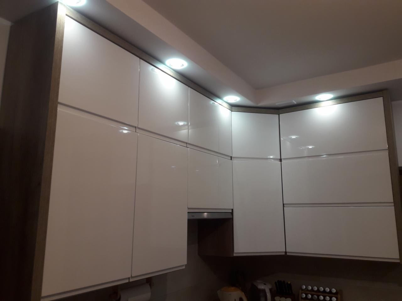 Białe, lakierowane szafki kuchenne podwieszane z drewnianymi obramowaniami, oświetlone punktowo, widoczne z dołu pod kątem.