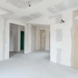 Wnętrze w trakcie prac wykończeniowych: ściany z płyt gipsowo-kartonowych, częściowo zagruntowane i zaszpachlowane, widoczne miejsca po wkrętach, surowa podłoga, instalacja elektryczna wyprowadzona...