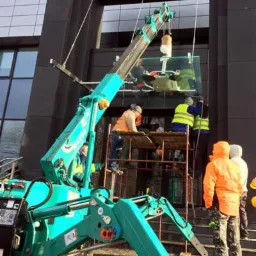 Montaż szklanej fasady biurowca Citi Handlowy z użyciem mini żurawia i rusztowania; pracownicy w odblaskowych kamizelkach nadzorują proces.