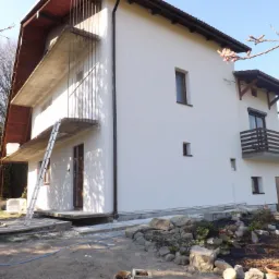 Dwukondygnacyjny dom z nową, białą elewacją, widoczne rusztowania i drabina, przygotowany teren wokół budynku z elementami skalniaka i roślinności.