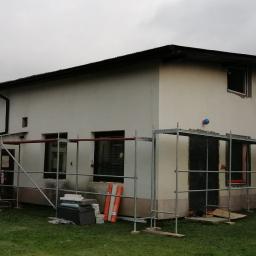 fasadapol - Dwukondygnacyjny dom w trakcie prac elewacyjnych, widoczne rusztowania, częściowo otynkowana ściana i materiały budowlane na trawniku, pochmurne niebo w tle.