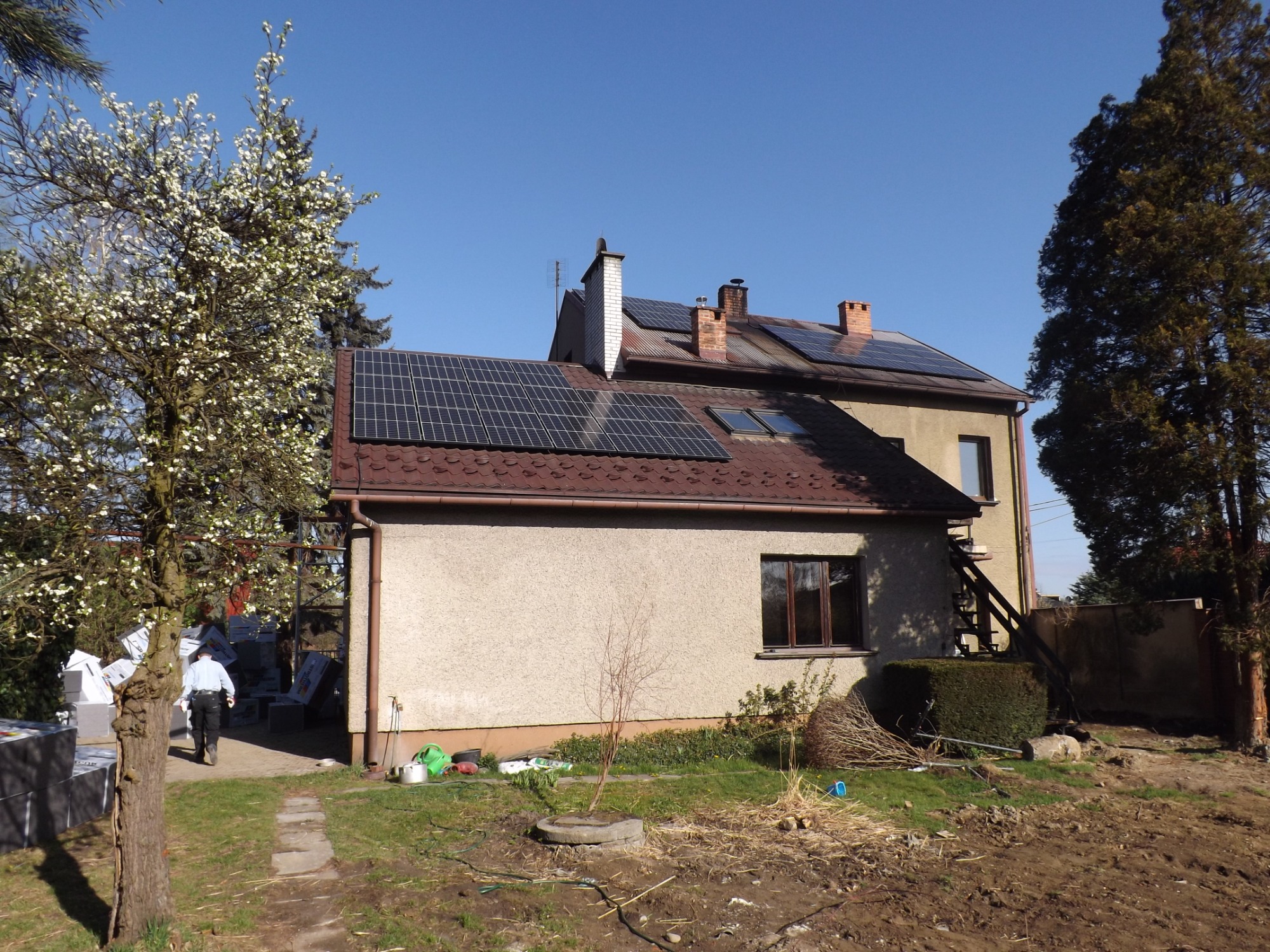 Dwukondygnacyjny dom z panelami słonecznymi na dachu, widok z ogrodu w słoneczny dzień, z widocznymi materiałami izolacyjnymi i pracownikiem w tle.