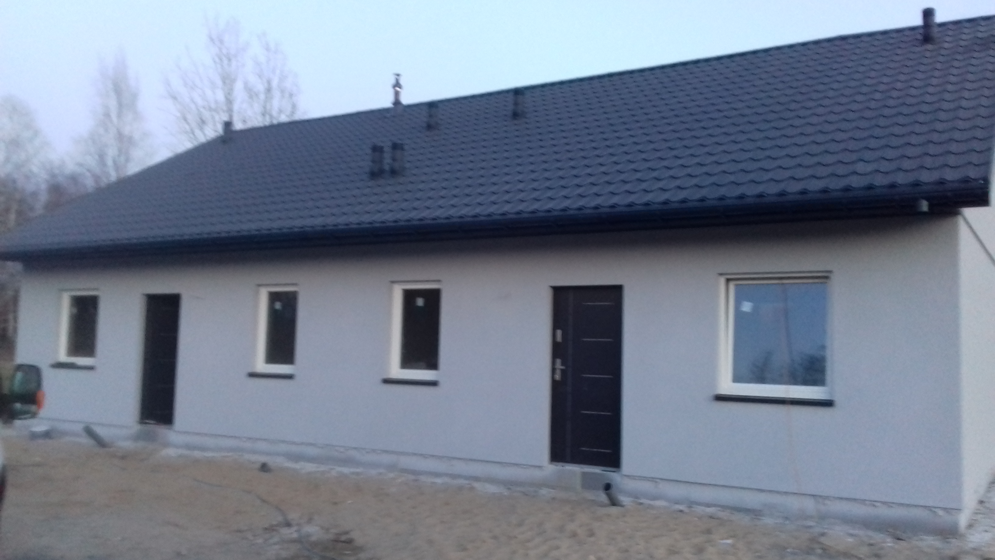 Nowo wybudowany parterowy budynek z szarą elewacją i ciemną dachówką, widoczne okna z białymi ramami i ciemne drzwi wejściowe, teren wokół w trakcie zagospodarowywania.