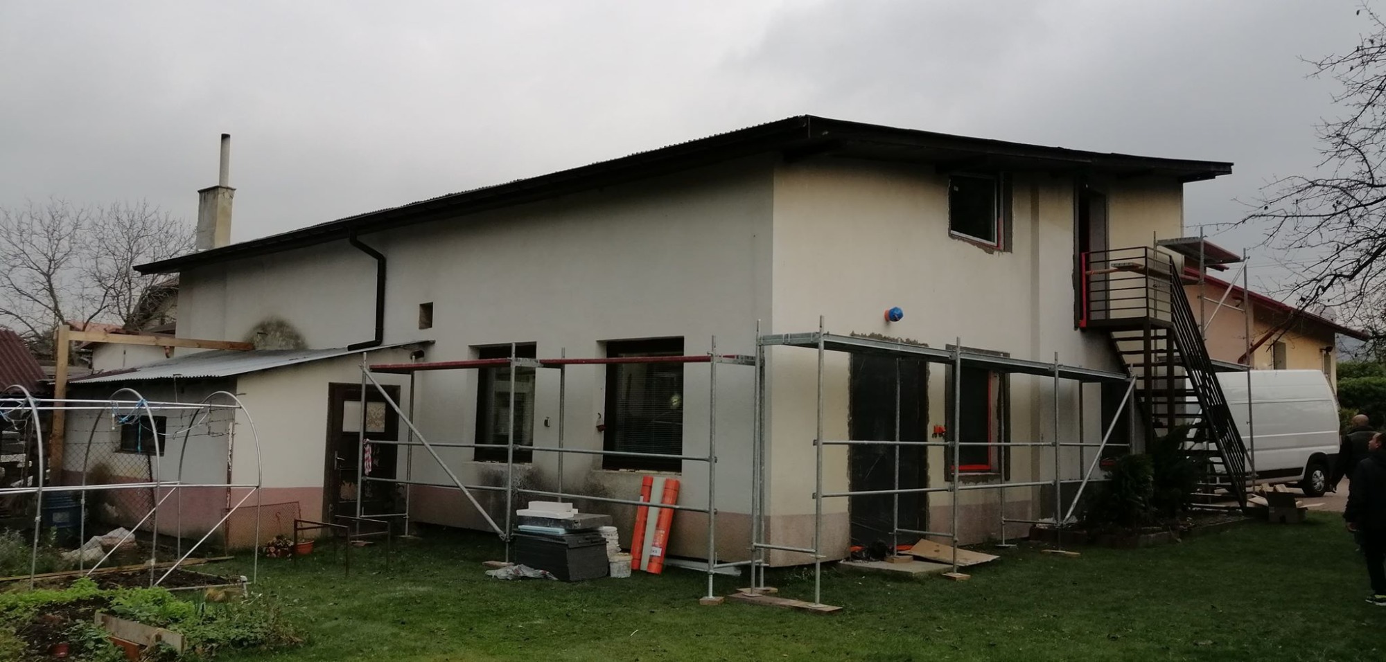 Dwukondygnacyjny dom w trakcie prac elewacyjnych, widoczne rusztowania, częściowo otynkowana ściana i materiały budowlane na trawniku, pochmurne niebo w tle.