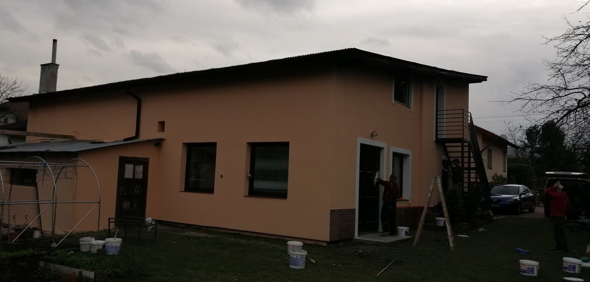 Dwukondygnacyjny dom z ocieploną elewacją w odcieniach beżu, z zewnętrznymi schodami, widocznymi oknami i drzwiami z białymi obramowaniami, pracownik malujący obramowanie drzwi, wiaderka z farbą...