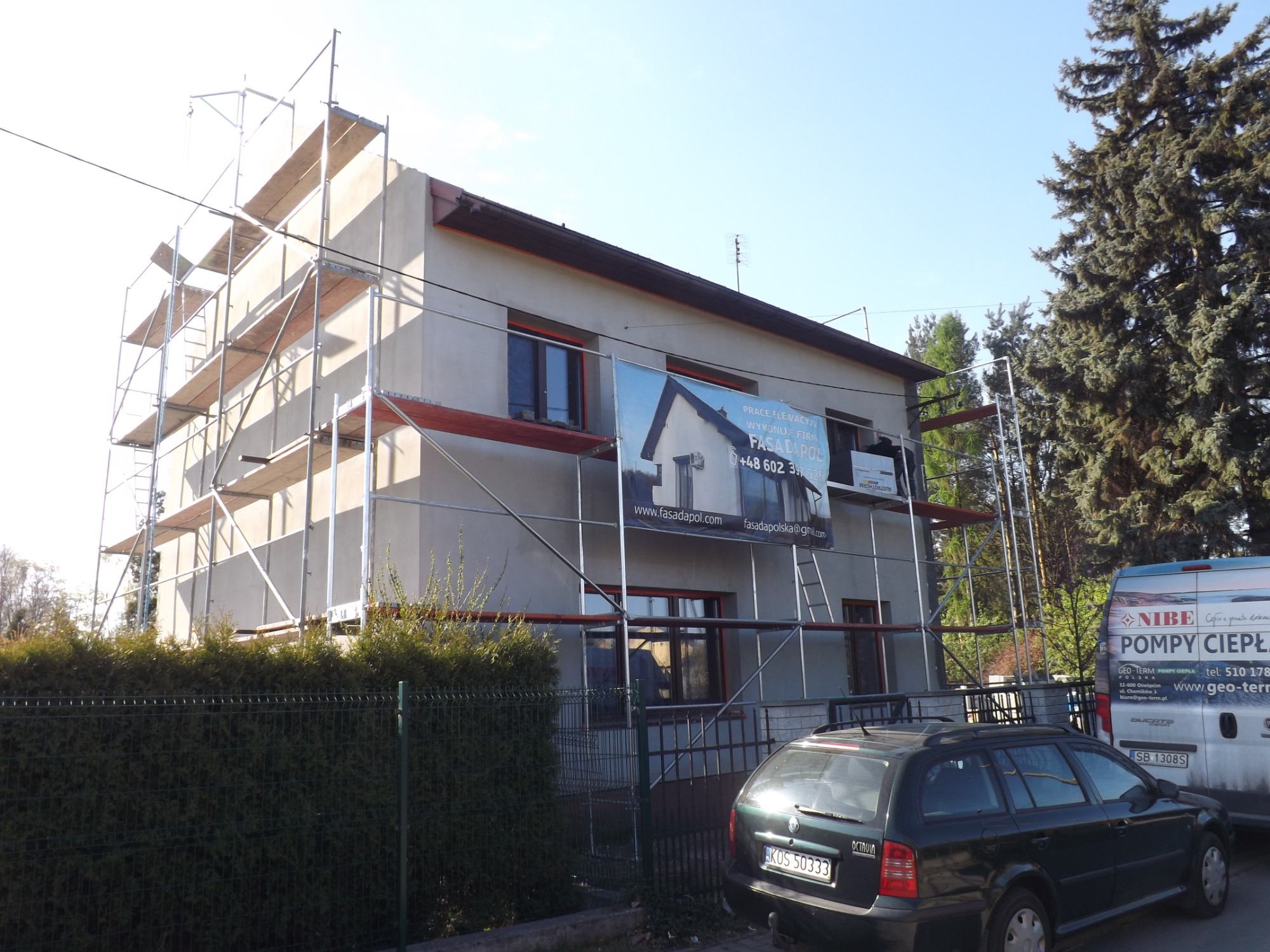 Dom jednorodzinny w trakcie prac elewacyjnych, otoczony rusztowaniem, z widoczną reklamą firmy wykonawczej i zaparkowanym samochodem osobowym oraz furgonetką z reklamą pomp ciepła.