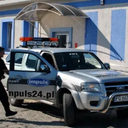 Mężczyzna w czarnym uniformie i kamizelce ochronnej otwiera drzwi srebrnego samochodu z napisem 'Monitoring' i logo firmy, zaparkowanego przed budynkiem z niebiesko-białymi pasami.