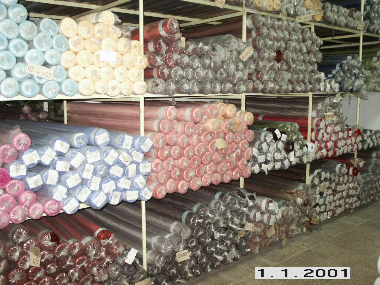 Magazyn z półkami pełnymi różnokolorowych bel materiałów, niektóre zapakowane w folię, z widocznymi etykietami z numerami.