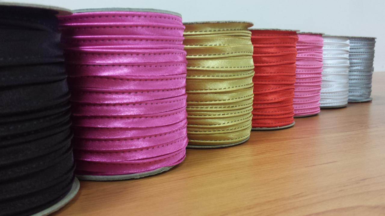 Szpulki satynowej lamówki w kolorach czarnym, różowym, złotym, czerwonym, jasnoróżowym, białym i srebrnym, ustawione w rzędzie na drewnianym blacie.