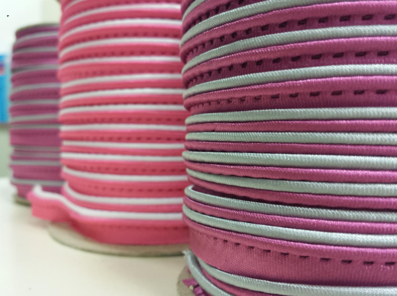 Szpule z różową i srebrną elastyczną lamówką, widoczne ściegi, ułożone w rzędzie na białym tle.