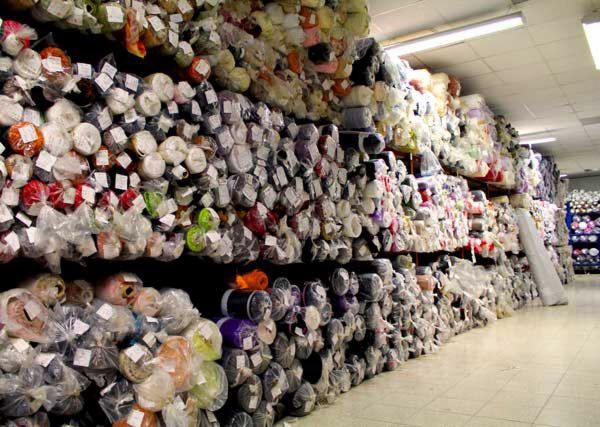 Magazyn pełen rolek tkanin w foliowych opakowaniach, ułożonych na regałach od podłogi do sufitu, z widocznymi etykietami na niektórych rolkach i jasnym oświetleniem sufitowym.