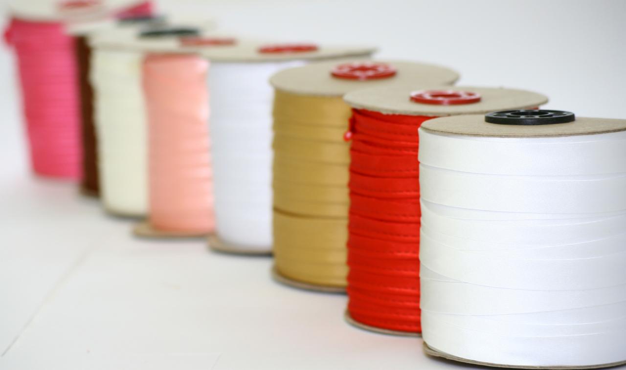 Rząd kolorowych wstążek satynowych nawiniętych na kartonowe szpule, z czarnymi i czerwonymi plastikowymi elementami mocującymi, ułożonych na białym tle.