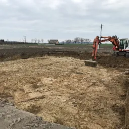 Pomarańczowa koparka Atlas 1604 wykonuje wykop pod fundamenty na tle pochmurnego nieba i odległego domu.