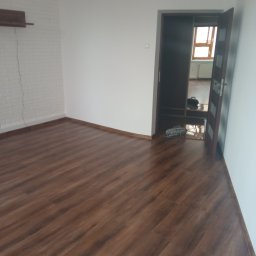 Krzysztof Kokosiński - Pusta przestrzeń po remoncie z nową, brązową podłogą z paneli, białe ściany, otwarte drzwi do sąsiedniego pomieszczenia z widocznym grzejnikiem i oknem.