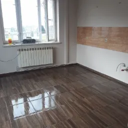 Wykończone wnętrze: ciemne, błyszczące płytki podłogowe imitujące drewno, częściowo wyłożona ściana brązowymi płytkami nad przyszłym blatem kuchennym, biały grzejnik pod oknem.