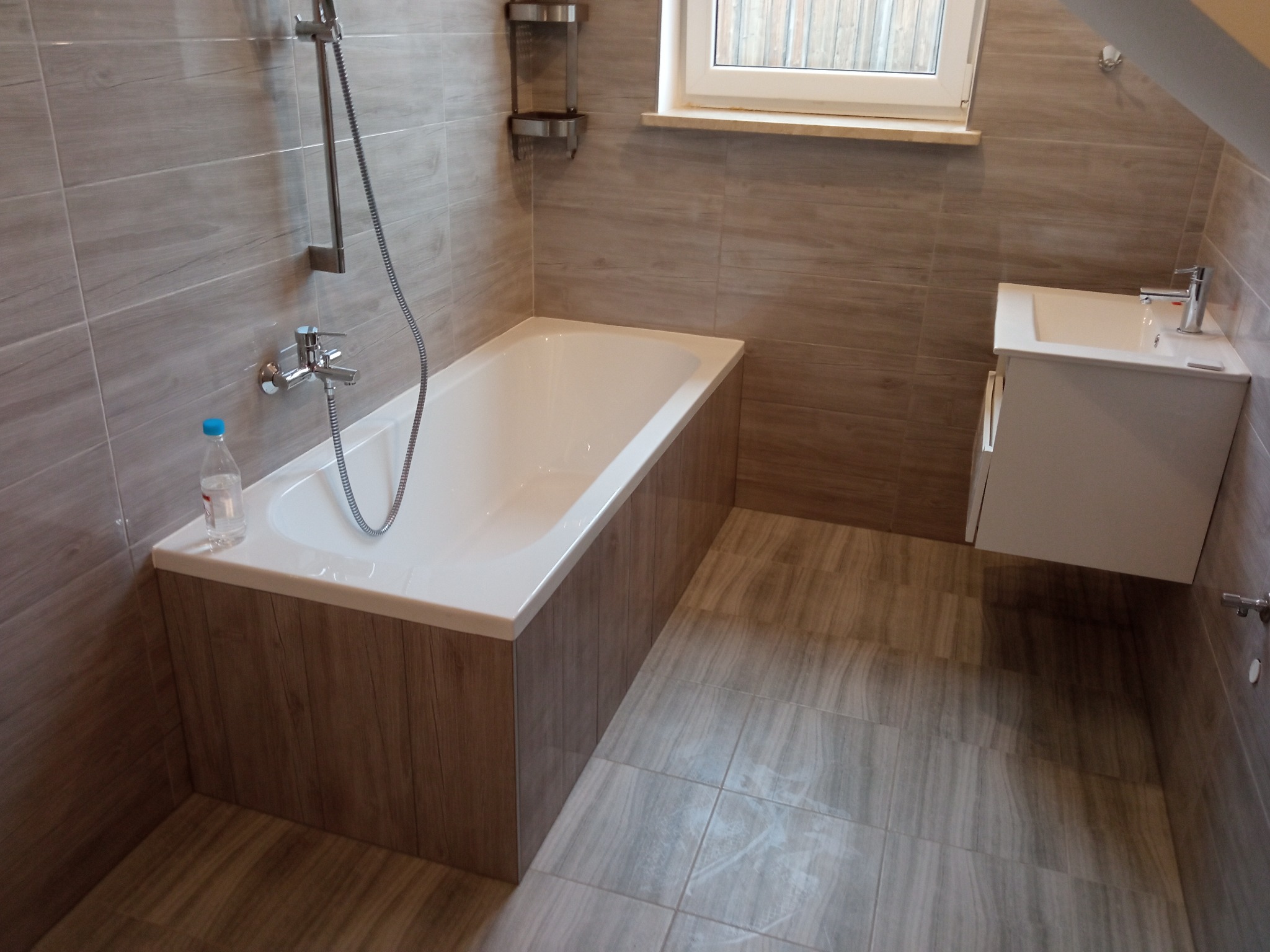 Nowoczesna łazienka z wanną obudowaną panelami imitującymi drewno, umywalką na szafce, szarymi płytkami imitującymi drewno na ścianach i podłodze, oraz butelką wody na brzegu wanny.