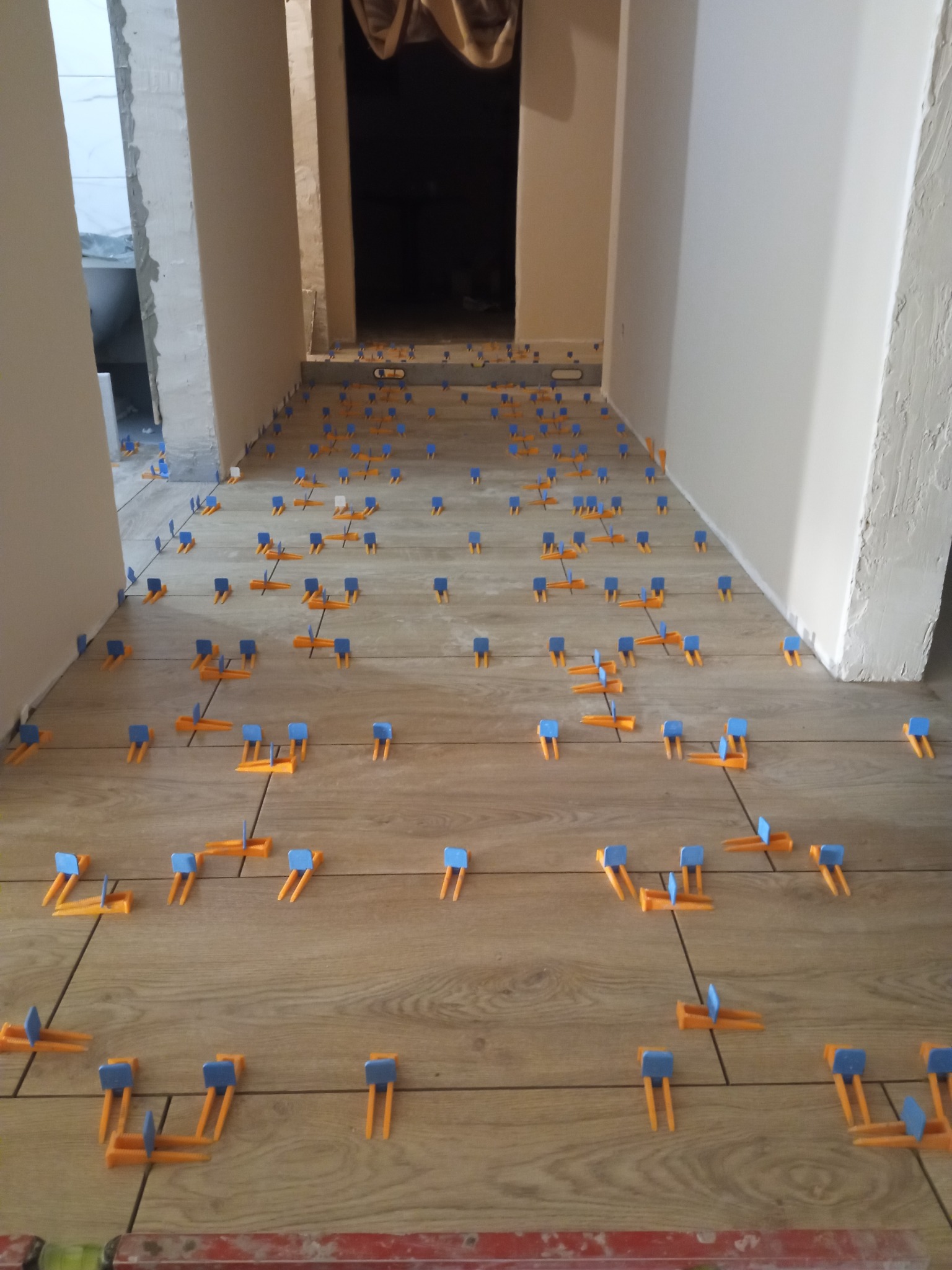 Układanie płytek podłogowych w korytarzu z użyciem pomarańczowo-niebieskich klipsów poziomujących, widok w głąb, ściany w stanie surowym.