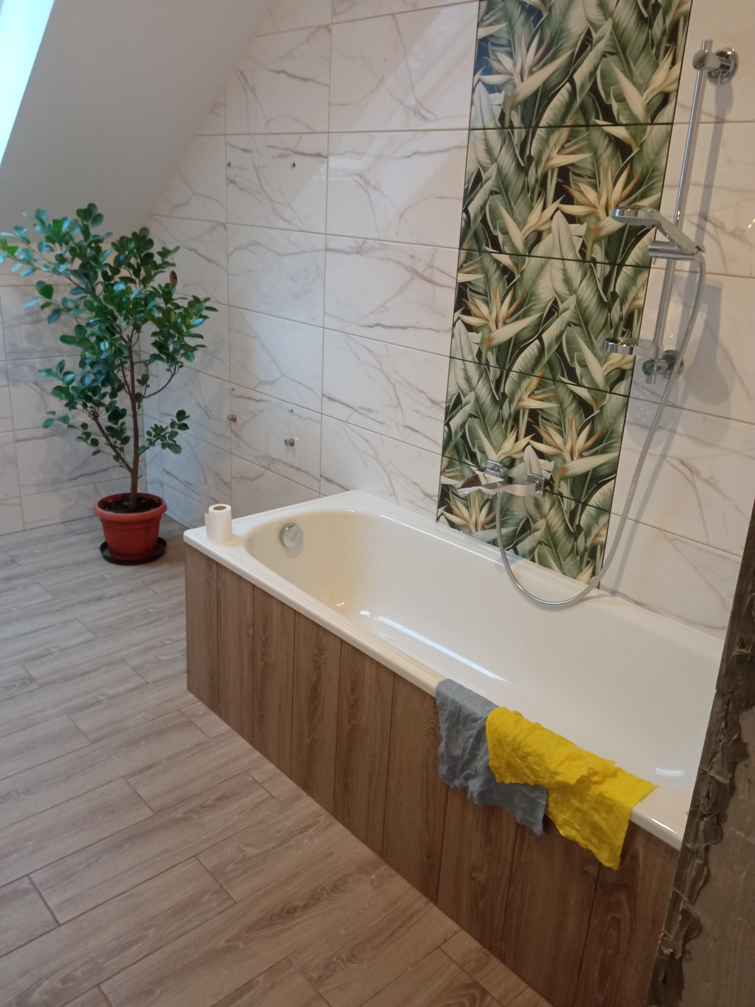 Wykończona łazienka na poddaszu z białą wanną obudowaną drewnopodobną okładziną, płytki imitujące marmur i z motywem roślinnym, armatura chromowana, na podłodze płytki imitujące drewno, obok wanny...