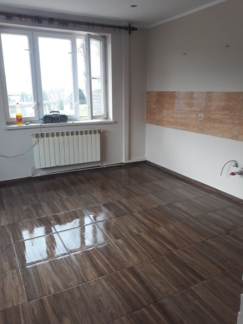 Wykończone wnętrze: ciemne, błyszczące płytki podłogowe imitujące drewno, częściowo wyłożona ściana brązowymi płytkami nad przyszłym blatem kuchennym, biały grzejnik pod oknem.