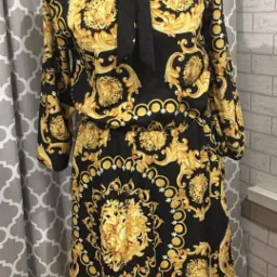 Czarno-złota sukienka w stylu barokowym z wiązanym kołnierzykiem, prezentowana na manekinie na tle szarej zasłony i ceglanej ściany.