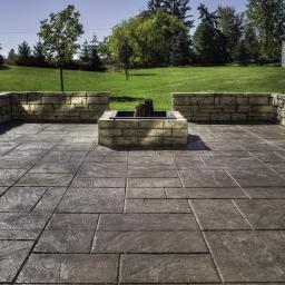 Fast Top Crete System - Zewnętrzna powierzchnia tarasu wykończona betonową posadzką z imitacją kamienia, otoczona niskim murem z jasnego kamienia i paleniskiem, na tle zielonego trawnika i drzew.