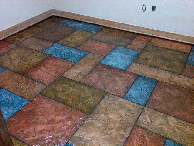 Wielobarwna posadzka z kwadratowych i prostokątnych płyt o zróżnicowanej fakturze, imitująca kamień w odcieniach brązu, czerwieni i błękitu, wykończona drewnianą listwą przypodłogową.