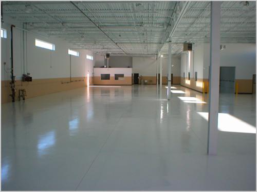 Biała, błyszcząca posadzka przemysłowa w dużej hali z widocznymi słupami i oknami w górnej części ścian.