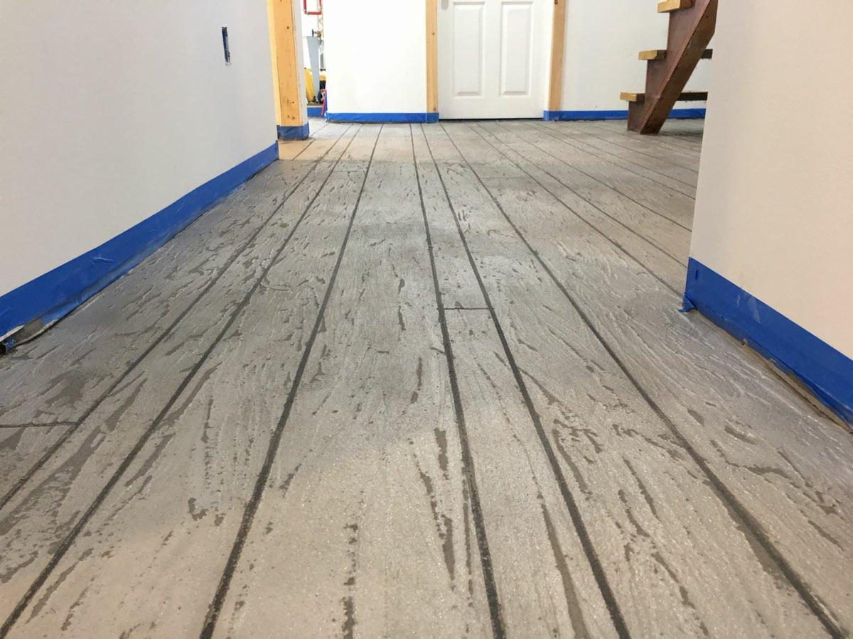 Szara podłoga imitująca drewniane deski, zabezpieczona niebieską taśmą malarską przy ścianach, z widocznymi schodami w tle.