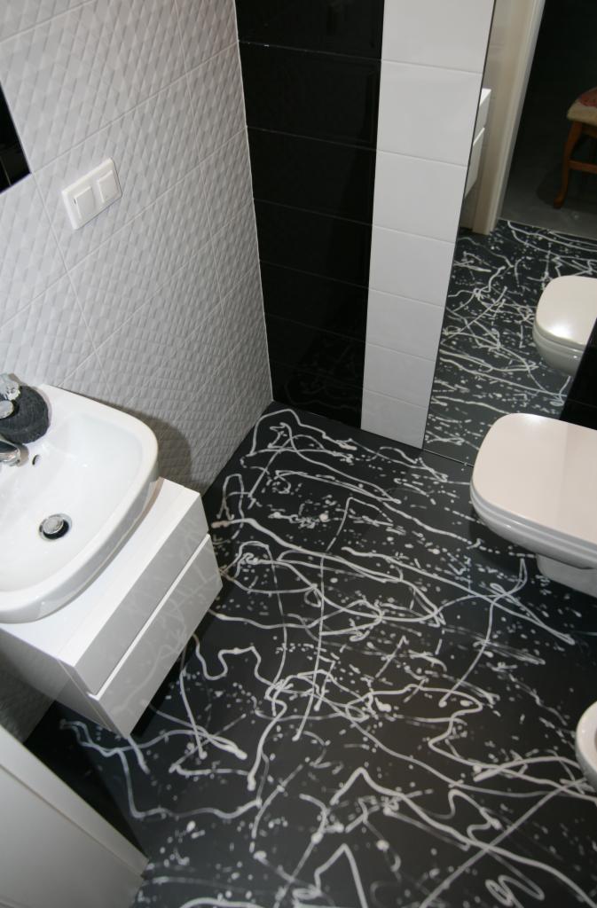 Czarna posadzka w łazience z białymi, nieregularnymi wzorami przypominającymi rozpryski farby, widoczna umywalka i toaleta w minimalistycznym stylu.