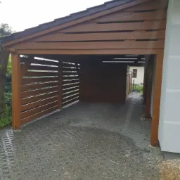 Zadaszone miejsce parkingowe z drewnianą konstrukcją i ażurowymi ścianami, wyłożone ażurową kostką brukową, widok z zewnątrz.