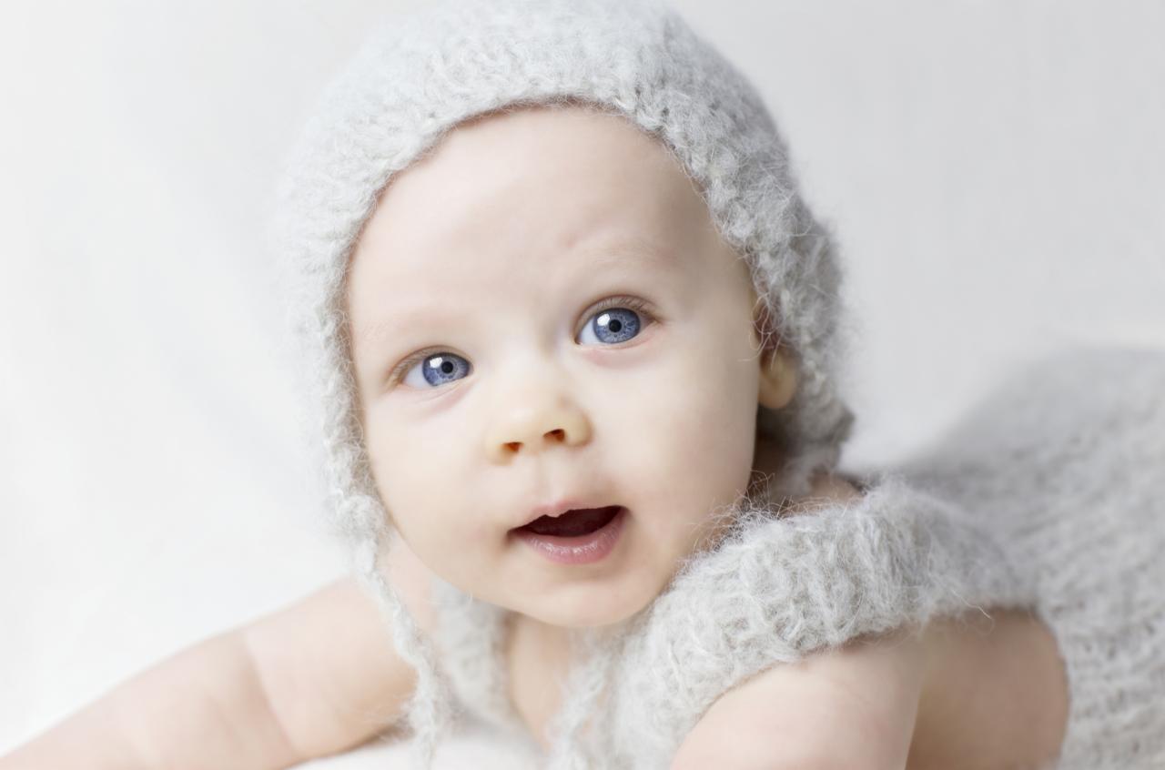 Niemowlę z otwartymi ustami, ubrane w futrzany kombinezon i czapeczkę, leżące na białym tle.