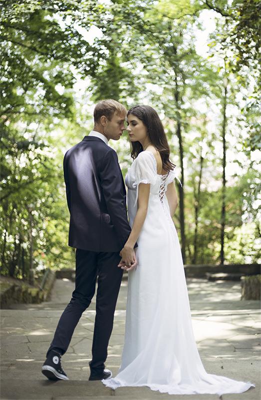 Para młoda trzymająca się za ręce na tle zieleni, on w garniturze i trampkach, ona w zwiewnej sukni z wiązaniem na plecach.