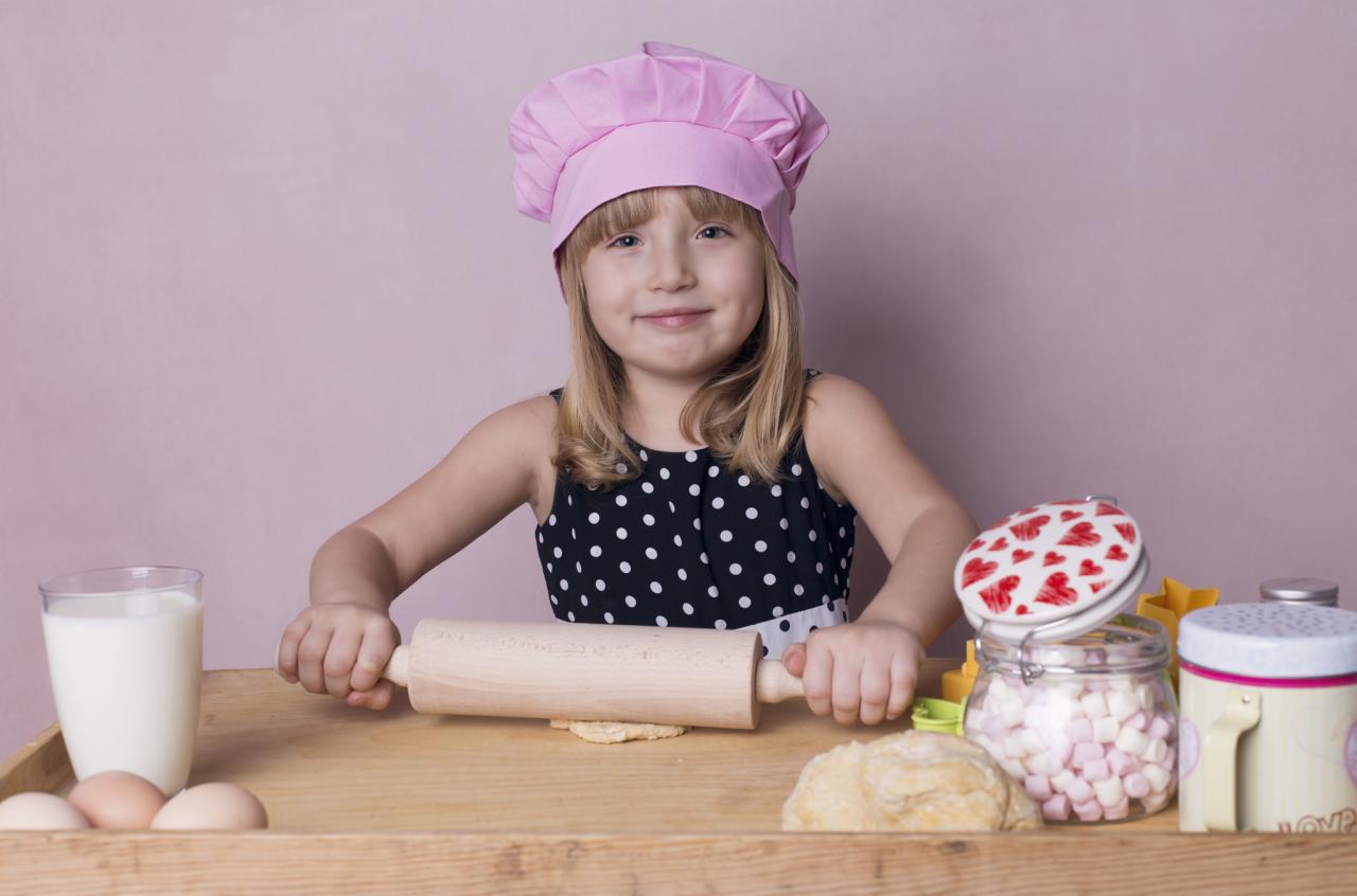 Dziewczynka w różowym czepku kucharskim wałkuje ciasto na drewnianym blacie, obok szklanka mleka, jajka, słoik z piankami i puszka z napisem LOVE na tle różowej ściany.