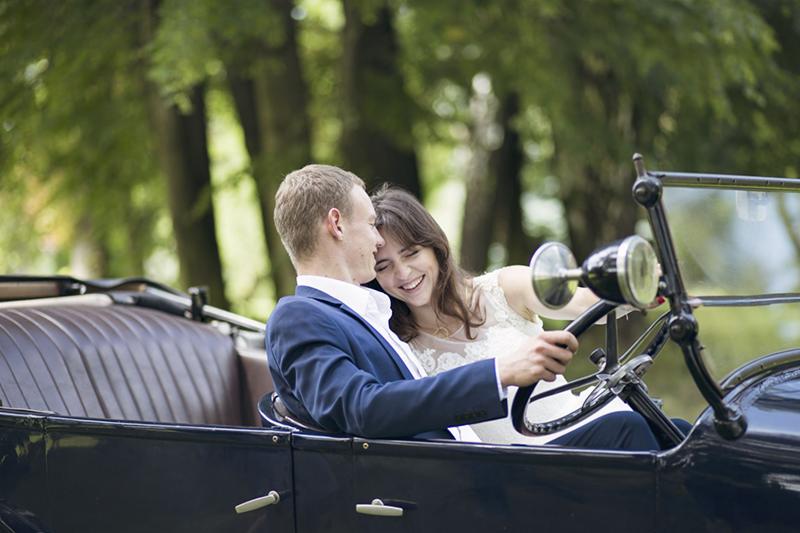 Uśmiechnięta para młoda w zabytkowym, czarnym kabriolecie na tle zieleni drzew.