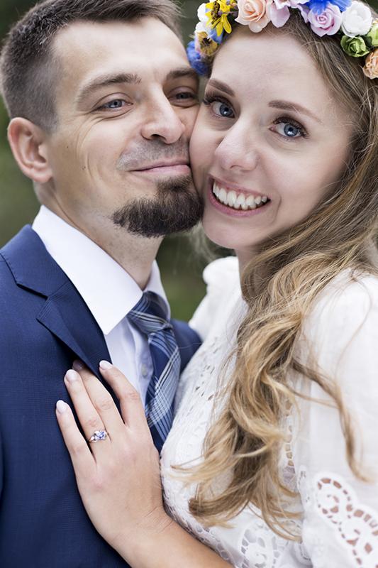 Uśmiechnięta para, zbliżenie na twarze i dłoń z pierścionkiem zaręczynowym; panna młoda ma wianek z kwiatów, pan młody w granatowym garniturze i krawacie w kratę.