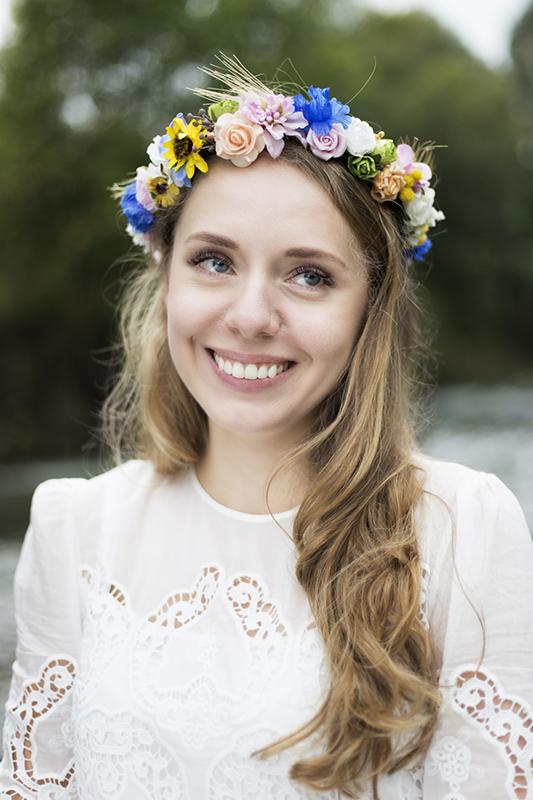 Portret uśmiechniętej młodej kobiety z wiankiem z kolorowych kwiatów na głowie, ubrana w białą koronkową bluzkę, z delikatnym makijażem i jasnymi włosami.
