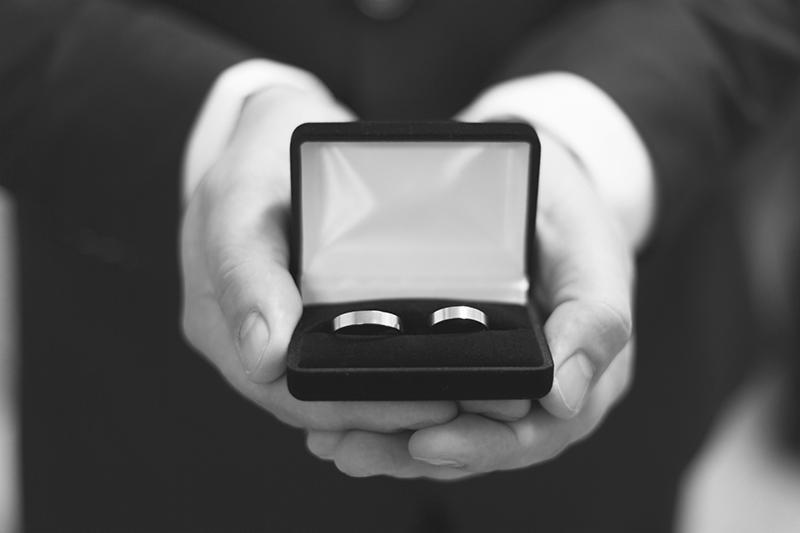 Czarno-białe zbliżenie dłoni mężczyzny w garniturze trzymających otwarte pudełko z dwoma gładkimi, srebrnymi obrączkami ślubnymi.