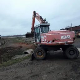 Czerwona koparka Atlas 1504 M na błotnistym terenie, rów z betonowymi rurami w tle, pochmurne niebo.